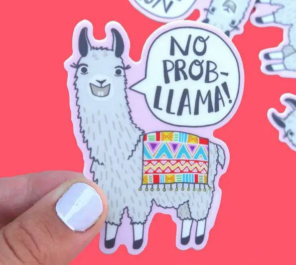 A hand with white nail polish holds a durable vinyl Llama No Prob-Llama Sticker by Lucky Sardine, featuring a smiling cartoon llama adorned with a colorful patterned blanket and a speech bubble saying "NO PROB-LLAMA!" The background is red, with other similar waterproof llama stickers faintly visible.