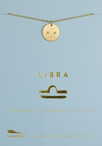 The LuckyFeather Zodiac Necklace Libra, featuring a gold round pendant with star and moon symbols, is elegantly displayed on a blue card. The card showcases "LIBRA" printed in gold, with the traits "ROMANTIC | CHARMING | PEACEFUL" listed below the zodiac sign symbol. It also mentions "14k gold dipped" and "adjustable.