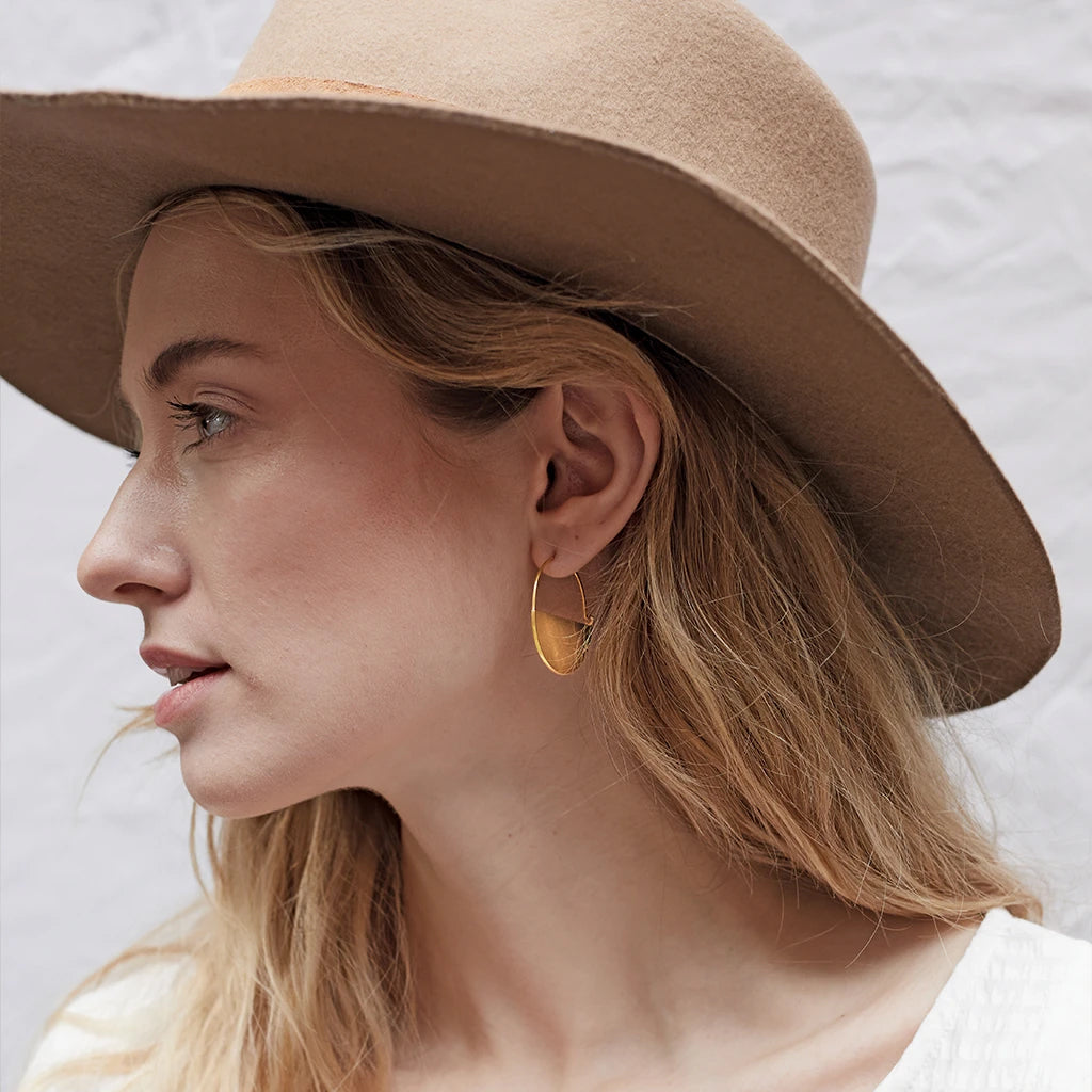 A person with long, light brown hair is wearing a wide-brimmed beige hat and a pair of Lunar Hoop Gold earrings from Scout Jewelry's Lightweight Refined Earring Collection. The person is shown in profile against a light-colored background, dressed in a white top.