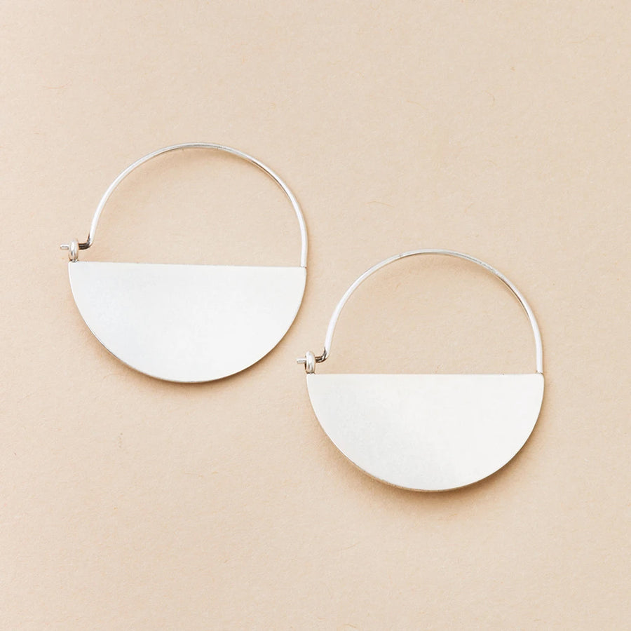 A pair of Lunar Hoop Silver earrings by Scout Jewelry is displayed on a beige background. Each hypoallergenic earring features a semi-circular, smooth sterling silver piece hanging from the bottom half of a thin, circular hoop.