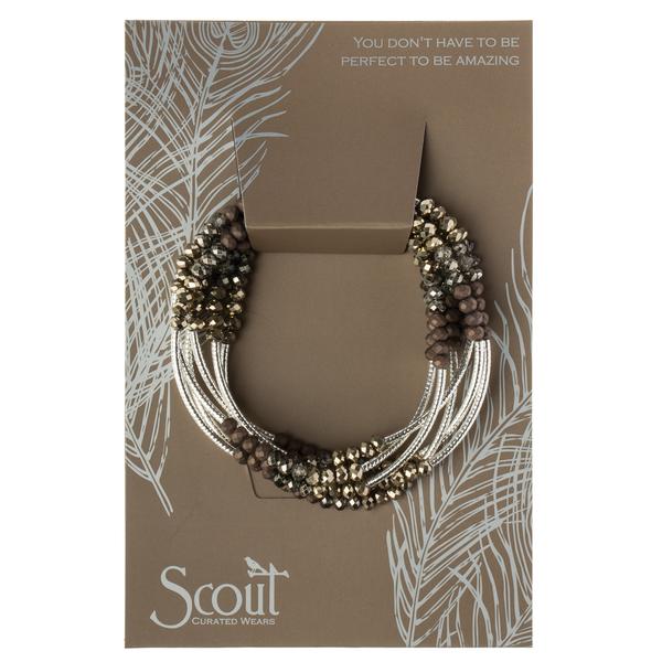 A necklace featuring multiple strands of crystal beads in metallic and earthy tones is showcased on a distinctive cardboard backing adorned with feather designs. The text includes, "Scout Curated Wears" and "You don't have to be perfect to be amazing." This piece, the Wrap Metallic Triple/Silver from Scout Jewelry, makes a thoughtful personalized gift or an inspiring addition as a Scout Wrap.