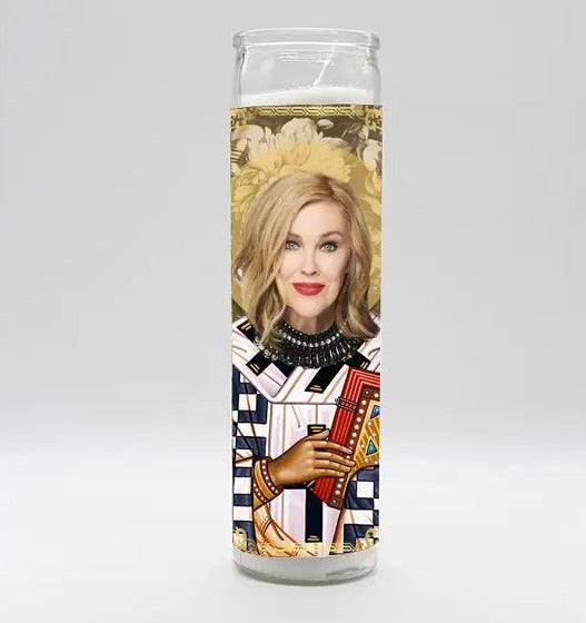 The Moira Rose Candle by BOBBYK BOUTIQUE is a tall, white candle emitting a gentle berry fragrance. It showcases an illustration of a woman with blonde hair and red lipstick in a majestic outfit, similar to a playing card design, set against detailed floral patterns.