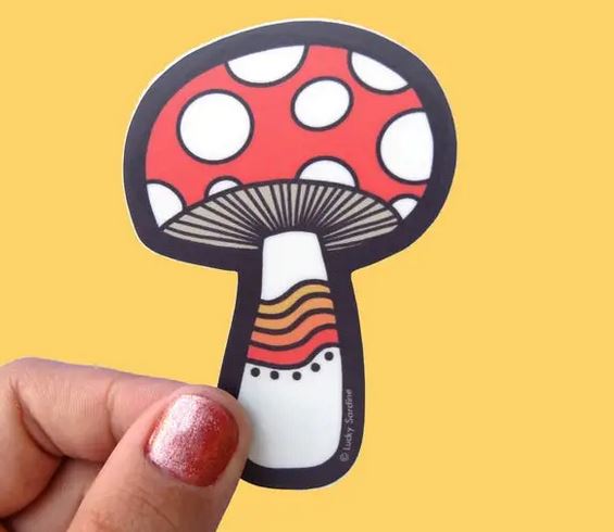 A hand with red glitter nail polish holds the Lucky Sardine "Mushroom Sticker," showcasing a scratch-resistant vinyl design featuring a red and white spotted mushroom with a black outline, set against a solid yellow background.