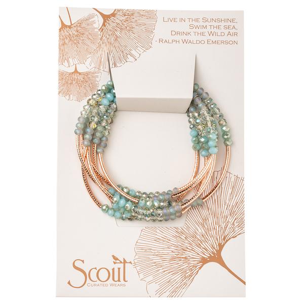 A multi-strand bead bracelet featuring crystal beads in blue and green stones is presented on a card with copper leaf designs. The card displays an inspiring quote by Ralph Waldo Emerson: "Live in the sunshine, swim the sea, drink the wild air." The brand "Scout Jewelry" is prominently visible.