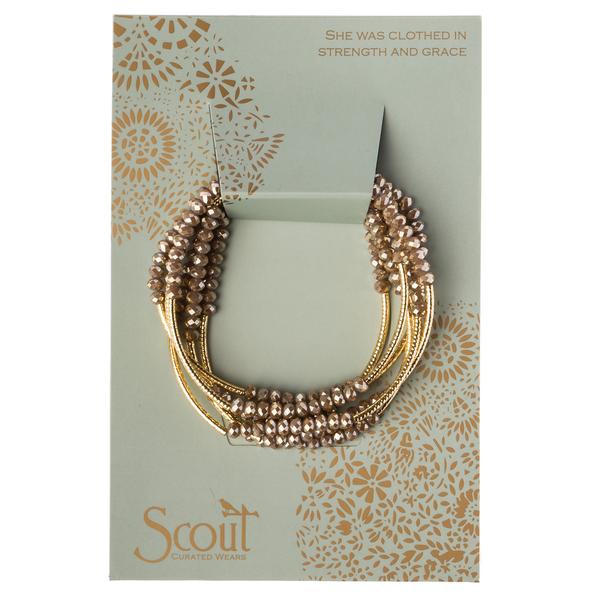 A set of golden beaded bracelets with crystal beads is presented on a light green decorative card adorned with floral patterns. The card features the phrase, "She was clothed in strength and grace," and is ideal for personalized gifts. The brand name, "Scout Jewelry," is elegantly printed at the bottom. This product is titled "Wrap Oyster/gold - She was clothed in.