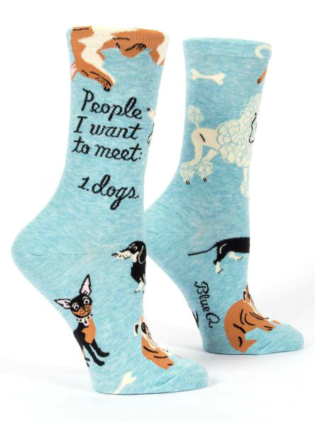 The People To Meet: Dogs Crew Socks by Blue Q are light blue cotton socks adorned with various dog illustrations, including a Chihuahua, Dachshund, Poodle, and others. One sock features black lettering that reads "People I want to meet: 1. dogs" and both women's shoe socks showcase the brand name "Blue Q" near the toes.