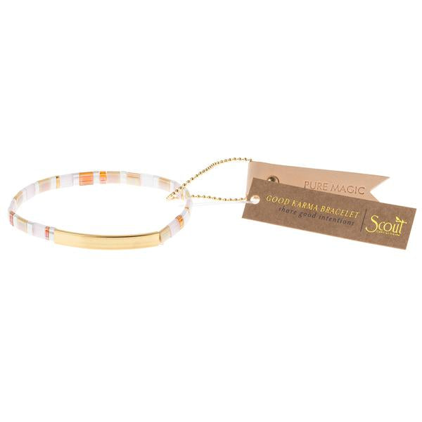 A delicate bracelet named "Miyuki Pure Magic Gold" by Scout Jewelry features a blend of gold, white, and orange beads with a 14K gold-dipped bar at the center. Attached is a brown tag inscribed with "PURE MAGIC" and "GOOD KARMA BRACELET" in gold text, accompanied by the Scout logo. The tag is affixed to the bracelet with a gold chain, radiating empowering messages.