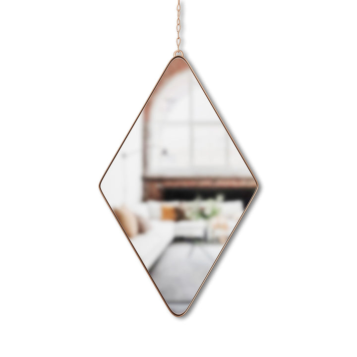 The Umbra Diamond Mirror Brass, featuring a gold frame and chain, elegantly reflects a cozy room with sofa, cushions, and large window, enhancing the warm atmosphere with its soft-focus charm.