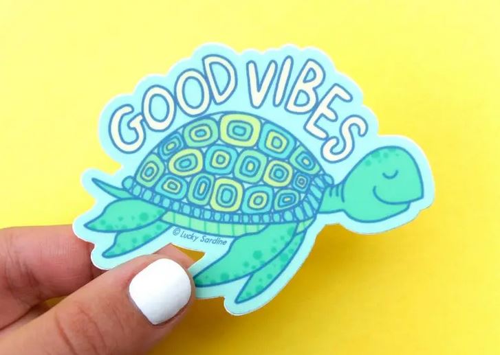 A hand with white nail polish holds the Sea Turtle Good Vibes Sticker by Lucky Sardine, which features a happy sea turtle with an intricately patterned shell. The words "GOOD VIBES" are written in bold, blue letters above the turtle, against a bright yellow background.