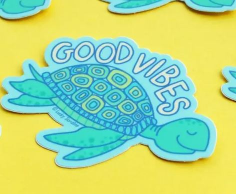 Introducing the Sea Turtle Good Vibes Sticker by Lucky Sardine: a cute, smiling sea turtle with intricate shell patterns set against a yellow background. The turtle's shell features the uplifting text "GOOD VIBES." This vibrant design in shades of green and blue is printed on weatherproof vinyl, ensuring it remains cheerful and bright regardless of the weather.