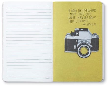 In the "Its Not What You Look At" notebook by COMPENDIUM, an open journal reveals a left page with blank lines and a right page with a vibrant yellow background, featuring an illustration of a camera alongside Joel Strasser's quote in elegant typography: "A good photographer must love life more than he does photography.