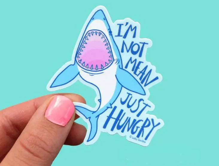 A hand with pink-painted nails holds a durable vinyl sticker from Lucky Sardine featuring a cartoon shark with its mouth wide open, revealing sharp teeth. The sticker, called "Shark I'm Not Mean Just Hungry Sticker," includes the text "I'M NOT MEAN JUST HUNGRY" on a solid light turquoise background.