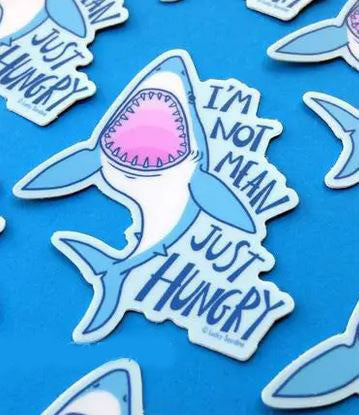 The Shark I'm Not Mean Just Hungry Sticker by Lucky Sardine features a playful illustration of a blue shark with its mouth open, revealing a pink inside, accompanied by the text "I'm not mean, just hungry" on a blue background. This whimsical design is surrounded by similar shark illustrations and is printed on durable, waterproof vinyl.