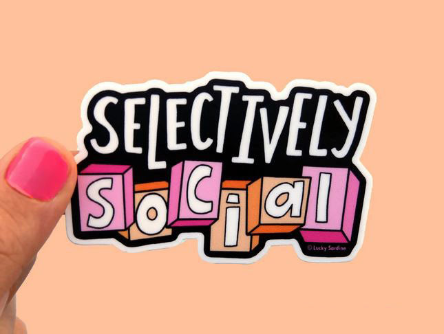 A hand with pink nail polish holds a weatherproof vinyl sticker from Lucky Sardine against a peach background. The Selectively Social Sticker, perfect for the funny introvert, features the phrase "Selectively Social" written on it. The word "Selectively" is in a bold, white font, and "social" is written in block letters with each letter in separate pink and orange boxes.