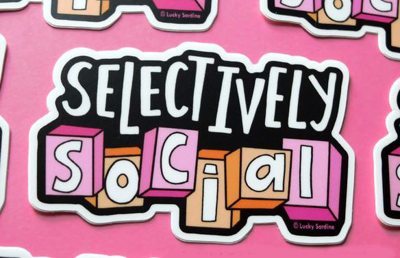 The **Selectively Social Sticker** by **Lucky Sardine** features the phrase "Selectively Social" in bold white text with a black outline. The word "Social" is displayed in block letters, each letter highlighted with shades of pink, purple, and orange, set against a pink background. It's the perfect funny introvert vinyl sticker for any occasion.