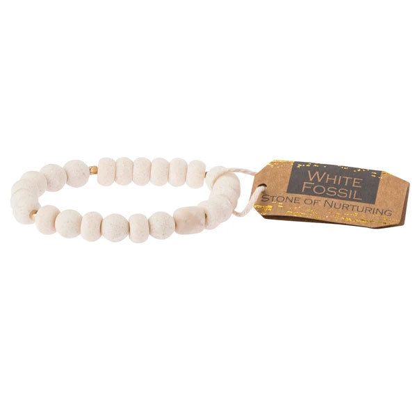 The White Fossil Stone Stacking Bracelet by Scout Jewelry showcases round white fossil beads with a unique look. A tag elegantly reads "White Fossil: Stone of Nurturing" in gold and black lettering, celebrating the semi-precious nature of this exquisite accessory.