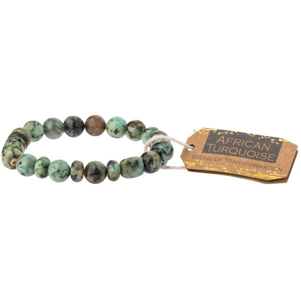 The African Turquoise Stone Stacking Bracelet by Scout Jewelry showcases turquoise-colored semi-precious gemstone beads in diverse green and brown shades. It includes a brown tag inscribed with "African Turquoise" and "Stone of Transformation" in gold lettering, bringing distinctive aesthetics and meanings.