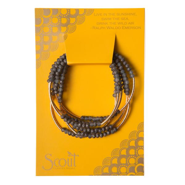 A beautifully arranged Scout Jewelry Wrap storm/gold bracelet set, adorned with dark beads and gold accents, is presented on an eye-catching yellow card. The card features a geometric design with an embossed quote by Ralph Waldo Emerson: "Live in the sunshine, swim the sea, drink the wild air," perfectly encapsulating the allure of this exceptional piece.