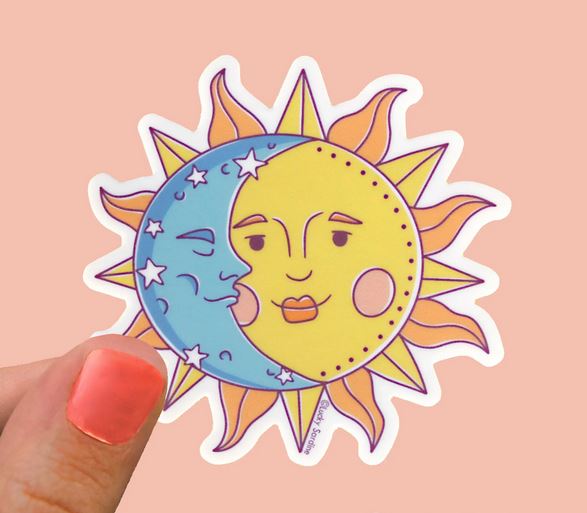 A person's hand with pink-painted nails holds the Sun and Moon Sticker by Lucky Sardine. The yellow sun, adorned with a content face and surrounded by rays, is partially overlapped by a serene-faced blue moon accented with stars. This weather-resistant sticker features a peach-colored background.