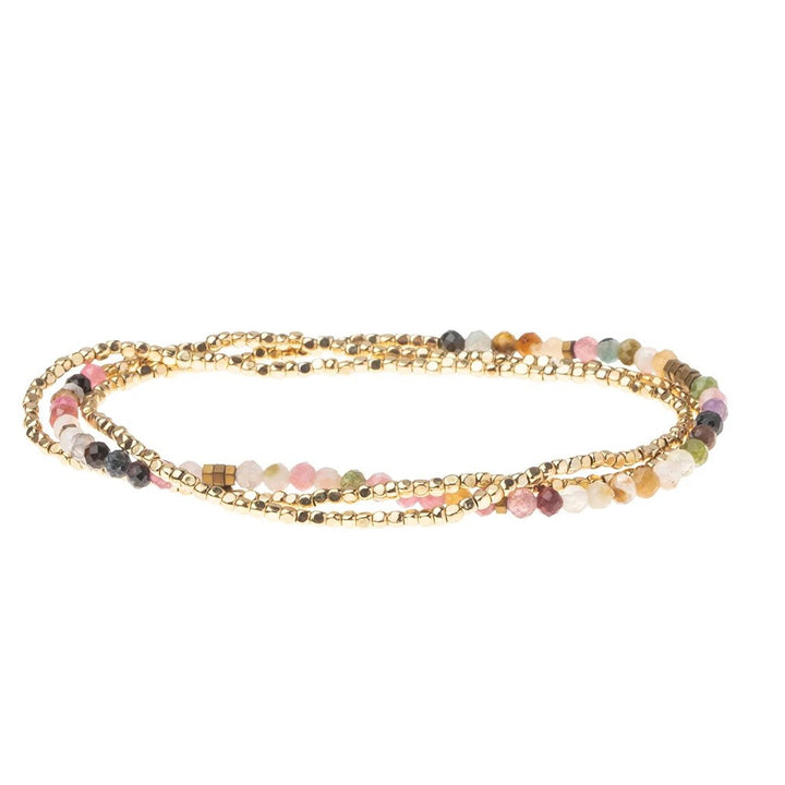 Introducing the Scout Jewelry Tourmaline Gold Delicate Wrap: a graceful, multi-strand bracelet showcasing an exquisite combination of small, colorful semi-precious tourmaline beads alongside lustrous gold-toned beads. This stunning gemstone bracelet is artfully arranged in a loose coil to highlight its vibrant mix of pink, green, white, and black beads interspersed with shimmering gold accents.