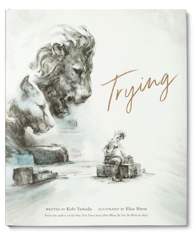 The book cover of the "Trying Book" by Kobi Yamada, published by Compendium, features a large statue of a contemplative lion alongside a smaller, possibly unfinished lion statue. A person sits nearby, drawing and expressing doubts while attempting something new. The title "Trying" is elegantly written in cursive.