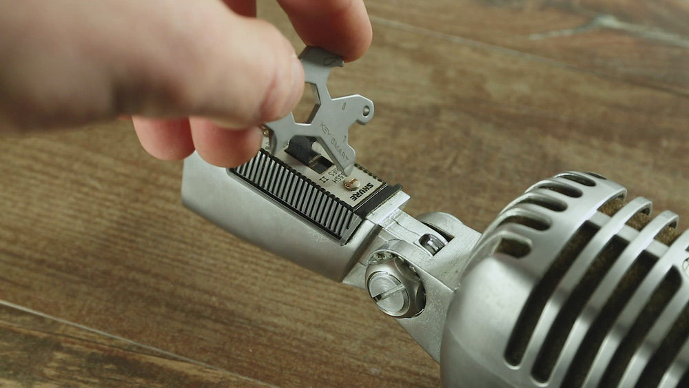 A hand is gripping the Dino Keychain Multitool by KeySmart, using it to adjust the screws on a vintage-style silver microphone sitting atop a wooden surface—an essential tool for the modern adventurer.