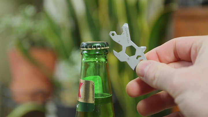 A hand holds the sleek and lightweight Dino Keychain Multitool by KeySmart, designed in silver and cleverly shaped like a key with various cutouts. It is poised above a green glass bottle, ready to skillfully open the cap. In the blurred background, indoor plants evoke an adventurer's spirit surrounding the scene.