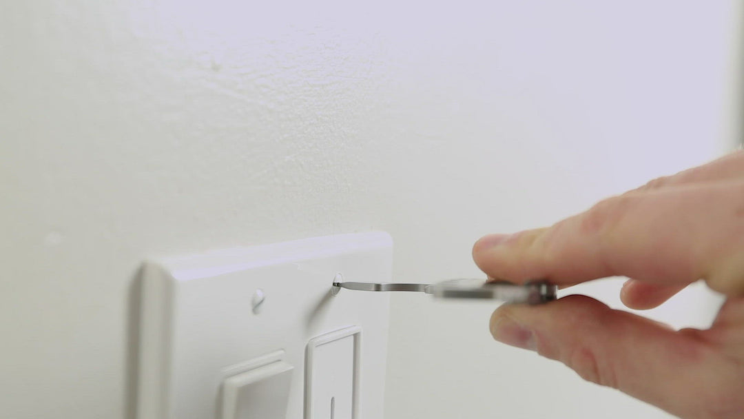 A close-up image of a hand holding the KeySmart Raptor Keychain Multitool, specifically using the flathead screwdriver feature, partially inserted into the screw of a light switch cover plate mounted on a neutral off-white wall. The action suggests someone is either tightening or loosening the screw.